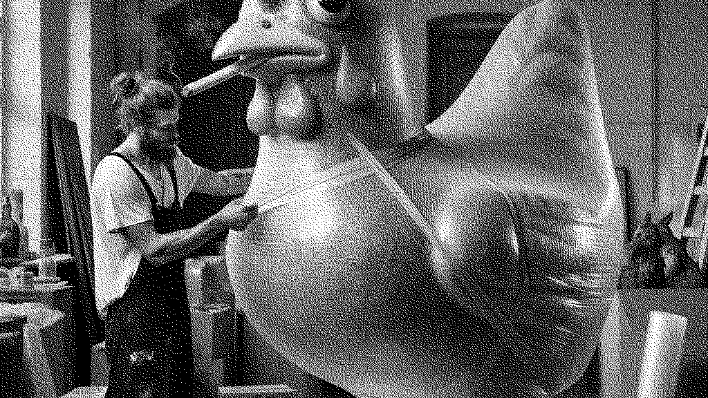 An art handler in overalls wrapping a scuplture of a chicken that is smoking a cigarette up in bubble wrap. The art handler looks like a hipster doofus with a man bun.